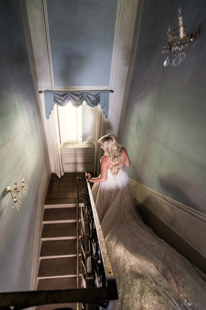 A bride in a white dress walks down a staircase, looking toward a window with blue curtains, before her Italian Villa wedding