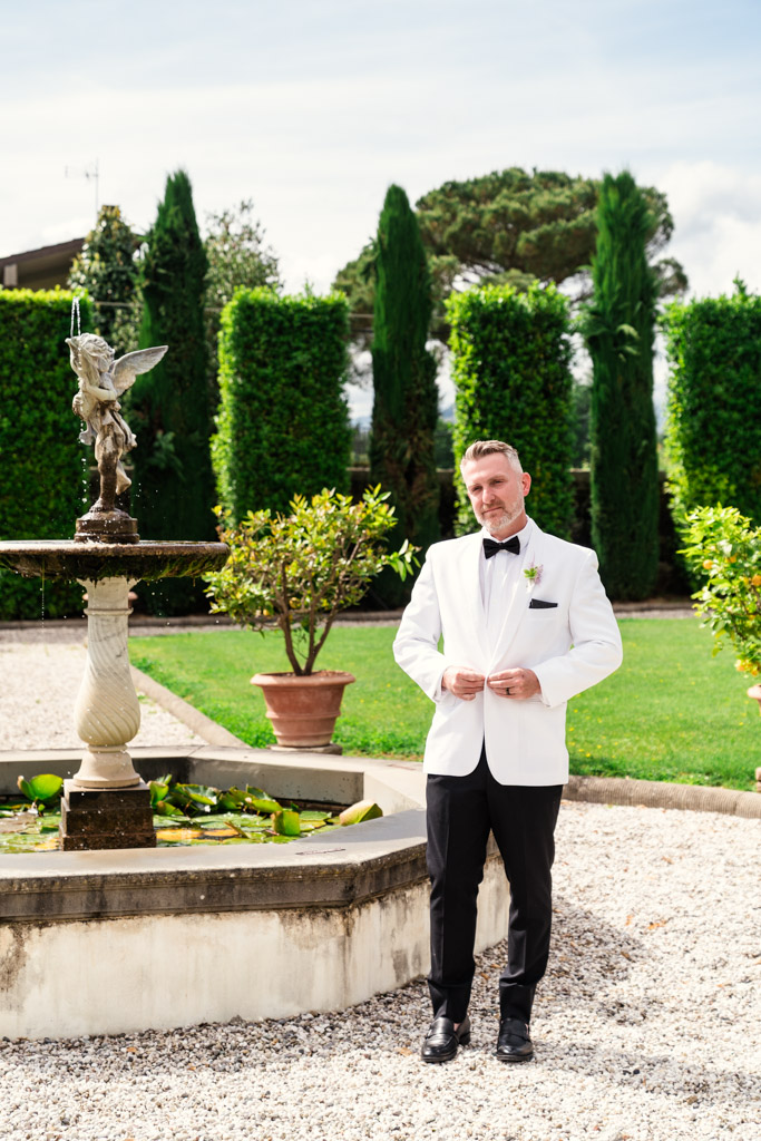Groom in white tuxedo stands by an Italian Villa garden fountain on a sunny day, with greenery in the background