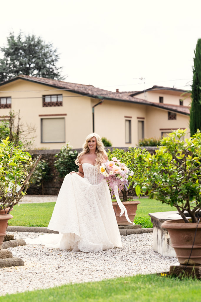 A bride in a white gown holds a large bouquet, walking outside among green plants and potted trees at her Italian Villa first look