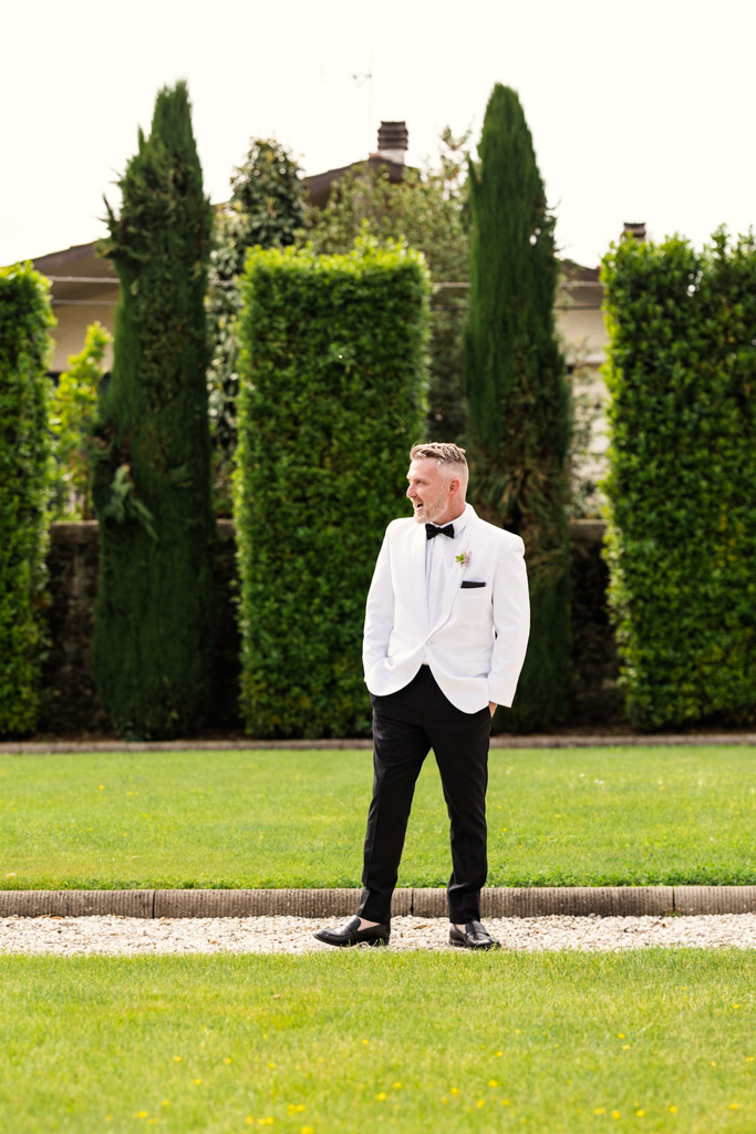 Groom in a white tuxedo jacket stands on a path in a garden with tall green hedges behind him, before his Italian Villa wedding