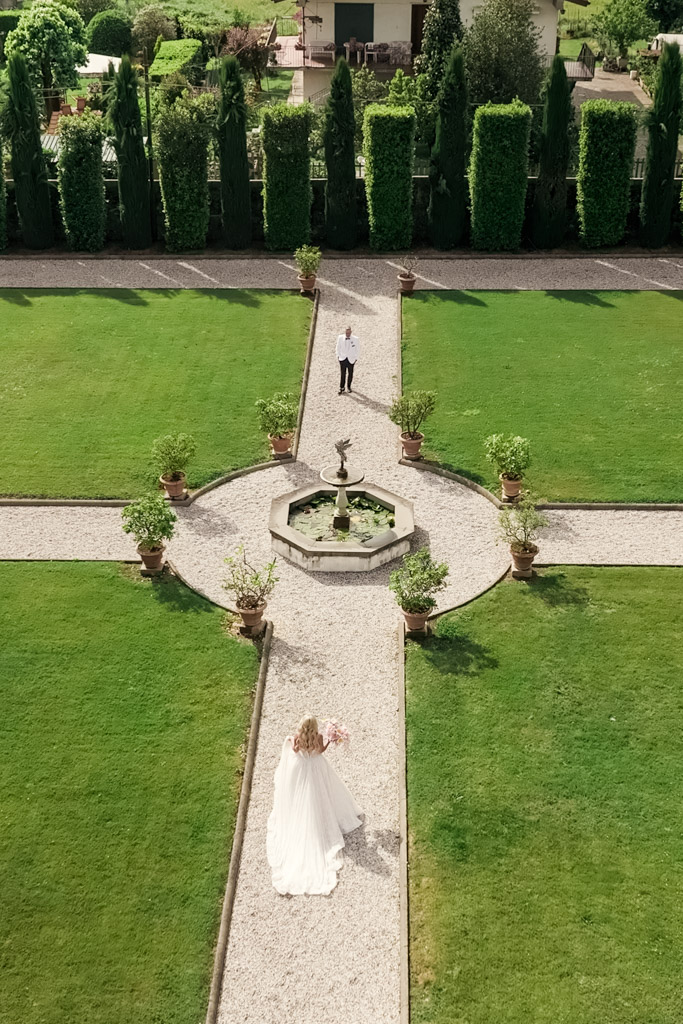 A bride in a white dress walks toward groom in a formal garden at an Italian Villa, paths forming a cross around a fountain