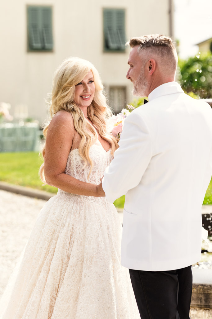 Bride and groom smile at each other outdoors during their Italian Villa wedding, bride in a white gown, groom in a white jacket, on a sunny day