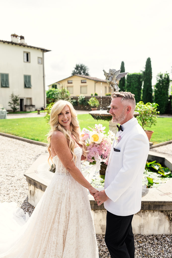 Bride and groom holding hands and smiling by a garden fountain on their Italian Villa wedding day