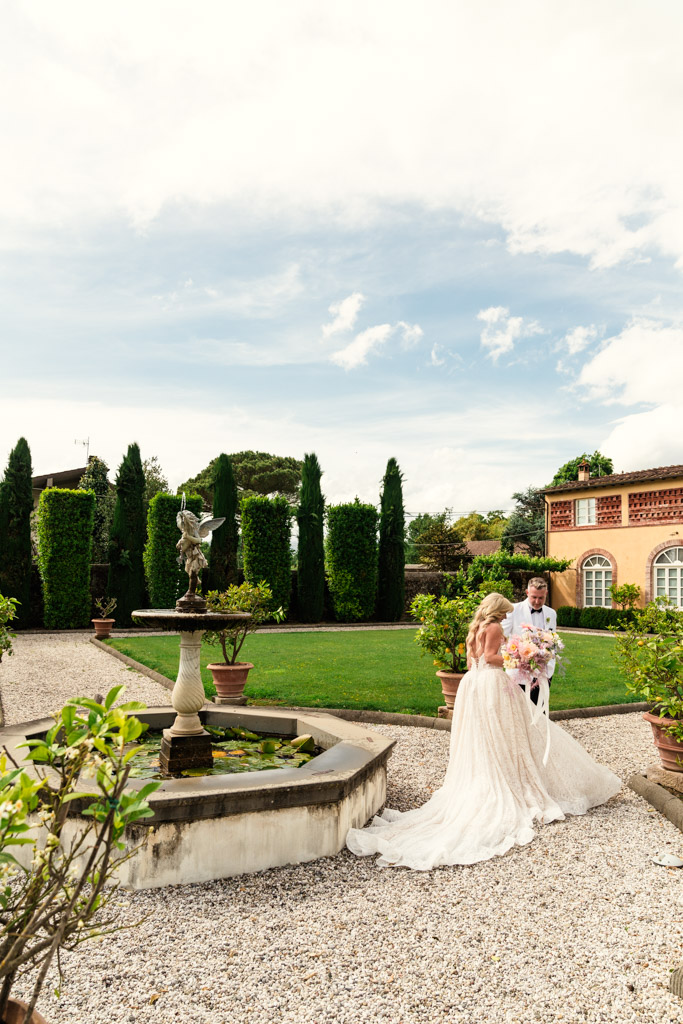 Bride and groom stand near a fountain in a garden, holding a bouquet, with lush greenery on the grounds of an Italian Villa