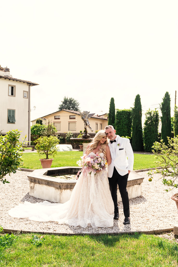 Bride and groom stand by a stone fountain in the garden of an Italian Villa wedding, smiling and dressed in elegant wedding attire