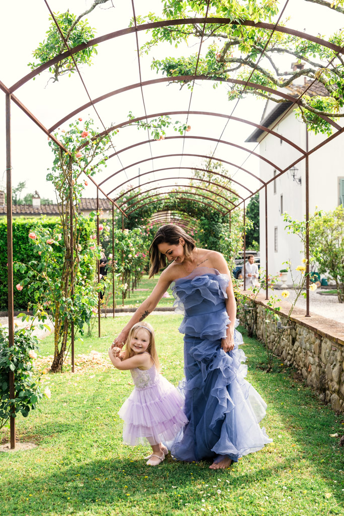 Woman in a blue ruffled gown and girl in a purple dress smiling together beneath a garden archway at Italian Villa wedding