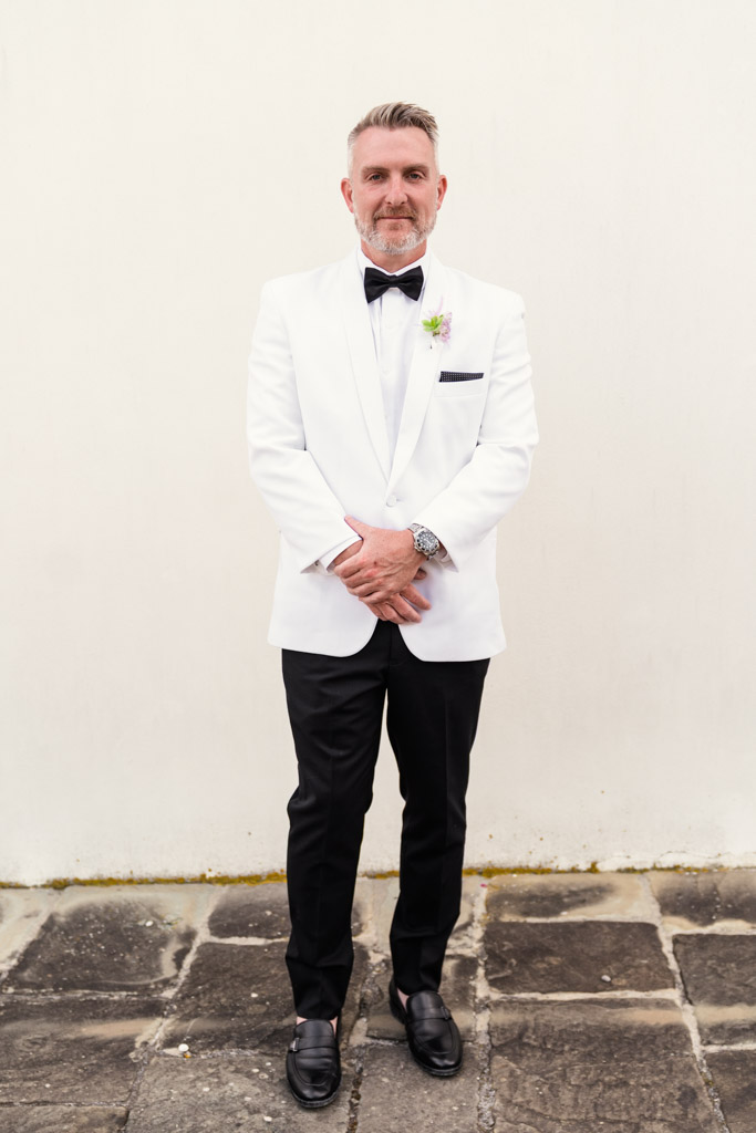 Portrait of groom in a white tuxedo jacket, black pants, and bow tie standing on stone pavement against a plain wall before his Italian Villa wedding