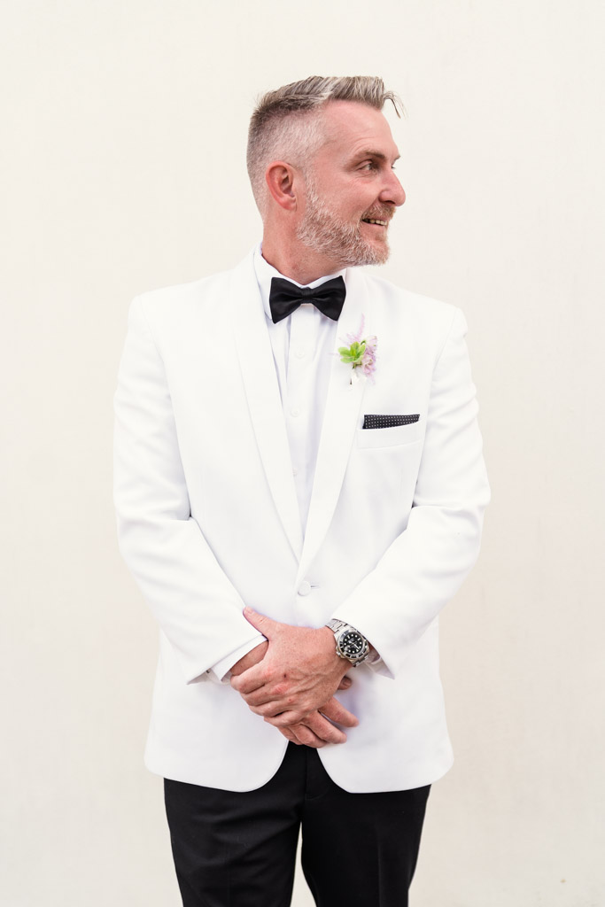Groom in a white tuxedo jacket and black bow tie stands smiling, looking to his right against a plain background before his Italian Villa wedding