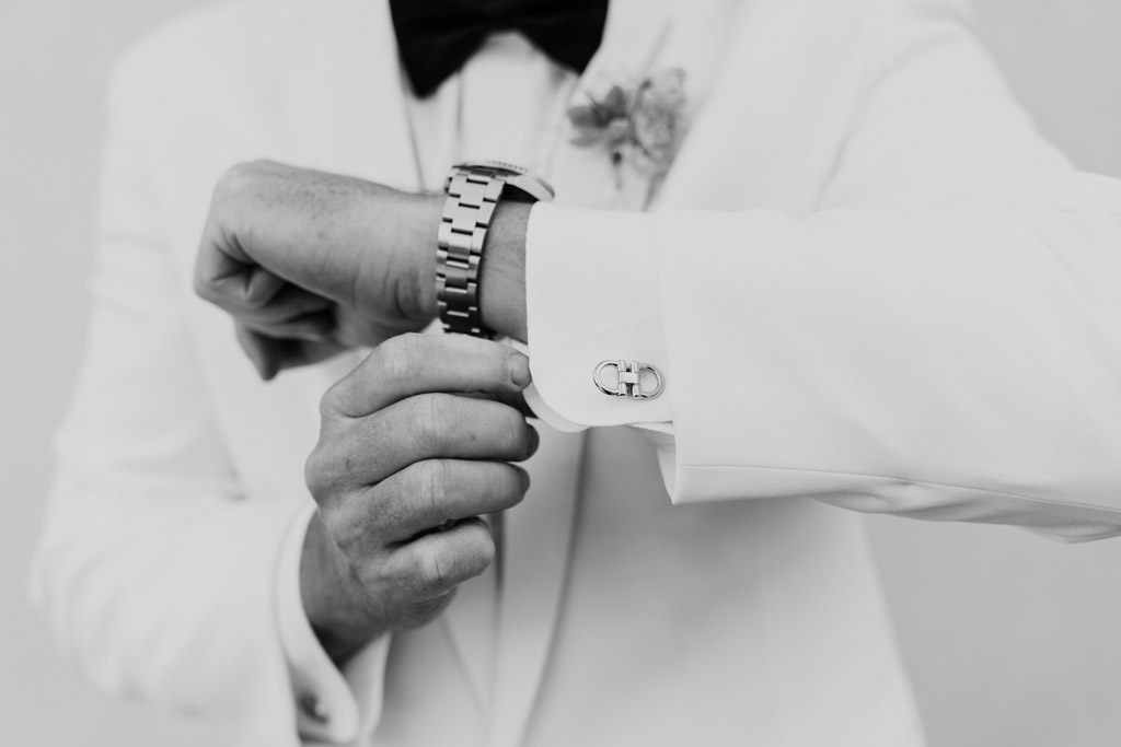 Black and white photo of groom in a white suit adjusting his cufflink and watch before his Italian Villa wedding; only his torso and arms are visible