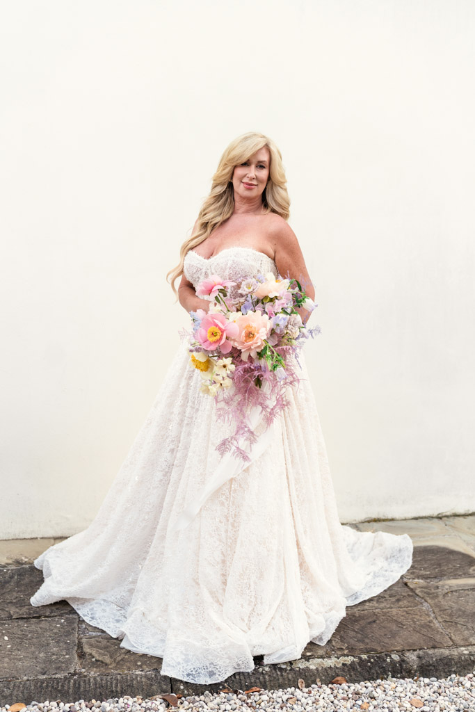Portrait of bride in a strapless white gown holds a colorful bouquet, standing on stone against a plain white wall before her Italian Villa wedding