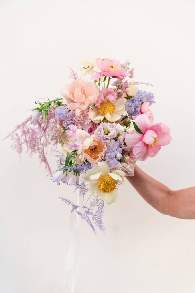 Bride's hand holds a bouquet of pastel flowers—including pink, yellow, and purple blooms against a white background