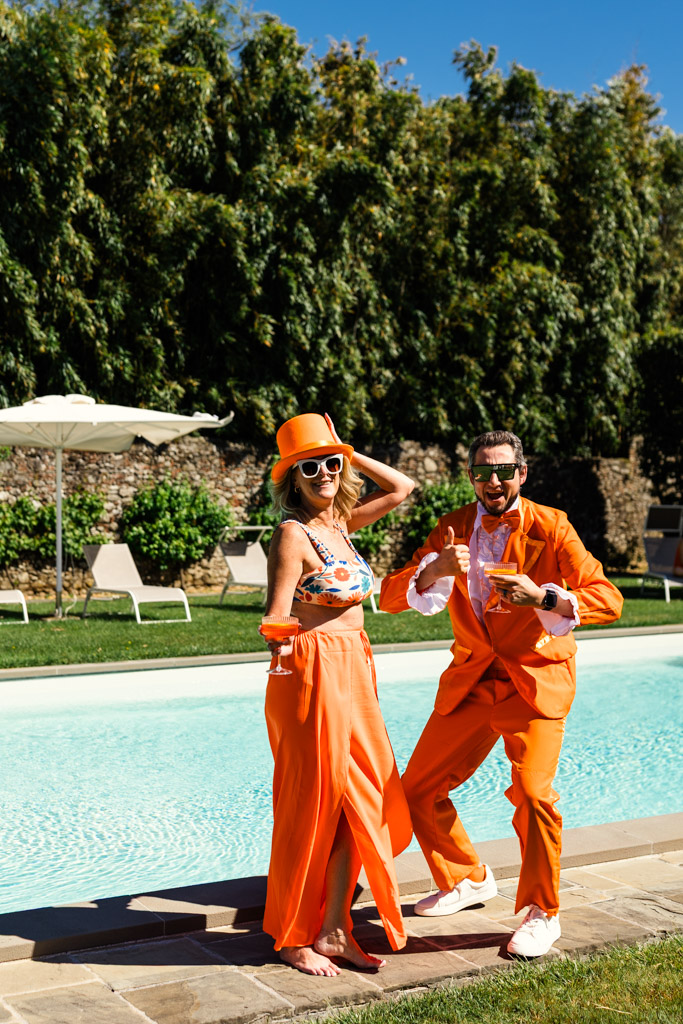 Two people in bright orange outfits pose playfully by a sunny outdoor pool with green trees in the background during post-wedding party at Villa Moscheni in Tuscany, Italy