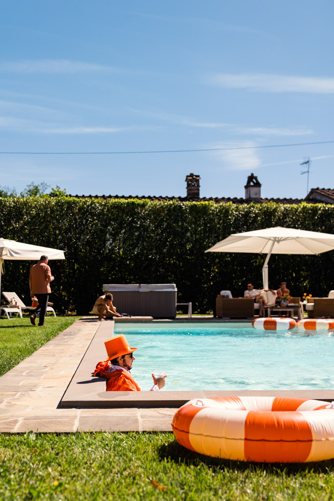 Person in orange hat and suit relaxes in a pool with a drink; others lounge nearby; lifebuoy on pool edge during Italy destination wedding pool party
