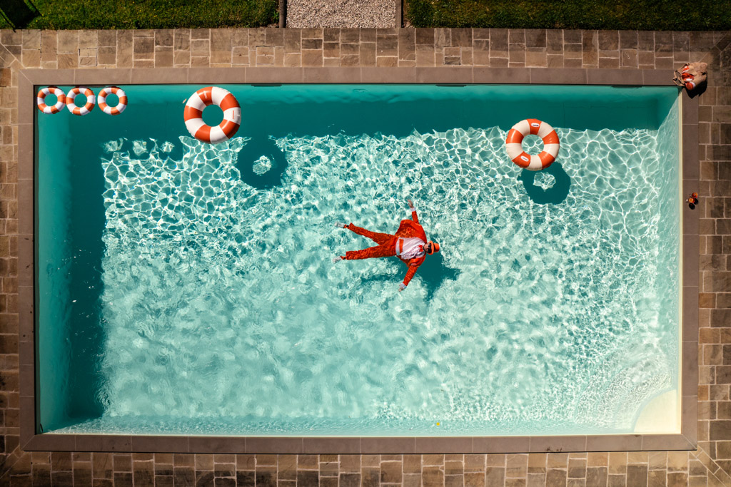 A person in an ornage suit floats in a pool surrounded by four life rings, viewed from above during wedding pool party at Villa Moscheni in Tuscany, Italy