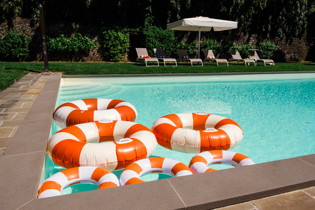 Six orange and white pool rings float in a clear swimming pool next to lounge chairs and a large umbrella during wedding pool party at Villa Moscheni in Tuscany, Italy