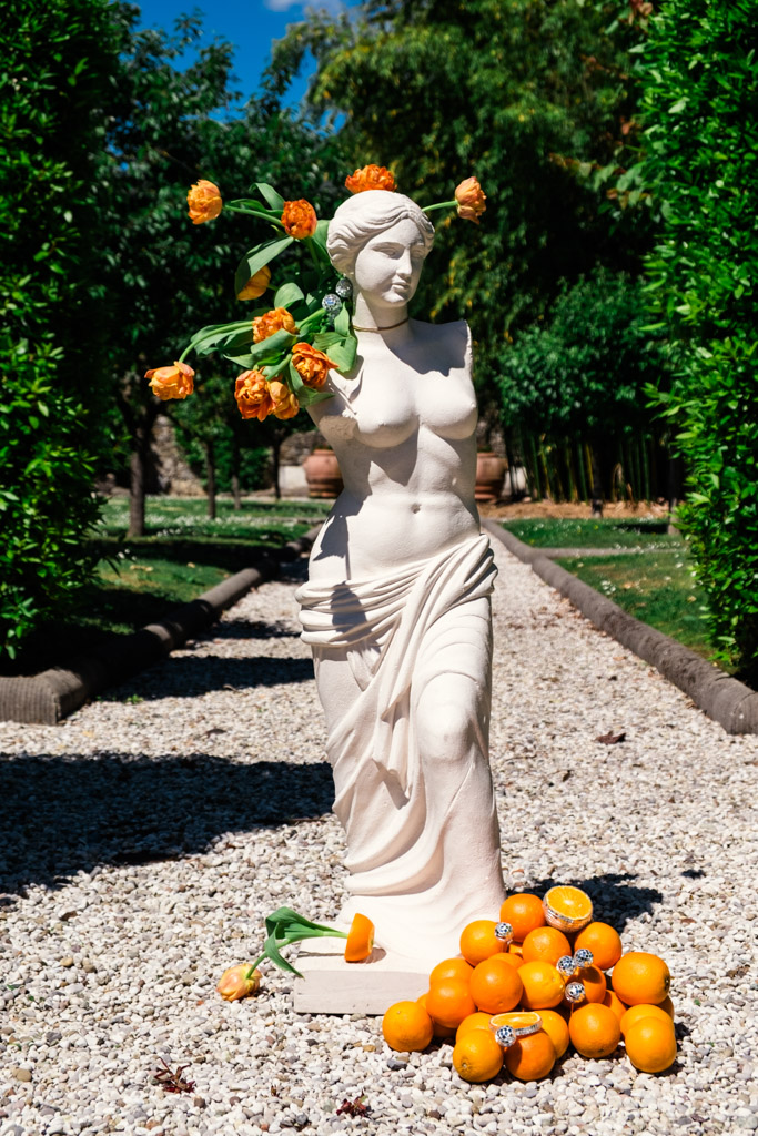 A marble statue with orange roses and scattered oranges stands on a gravel path in a garden at Villa Moscheni in Tuscany, Italy