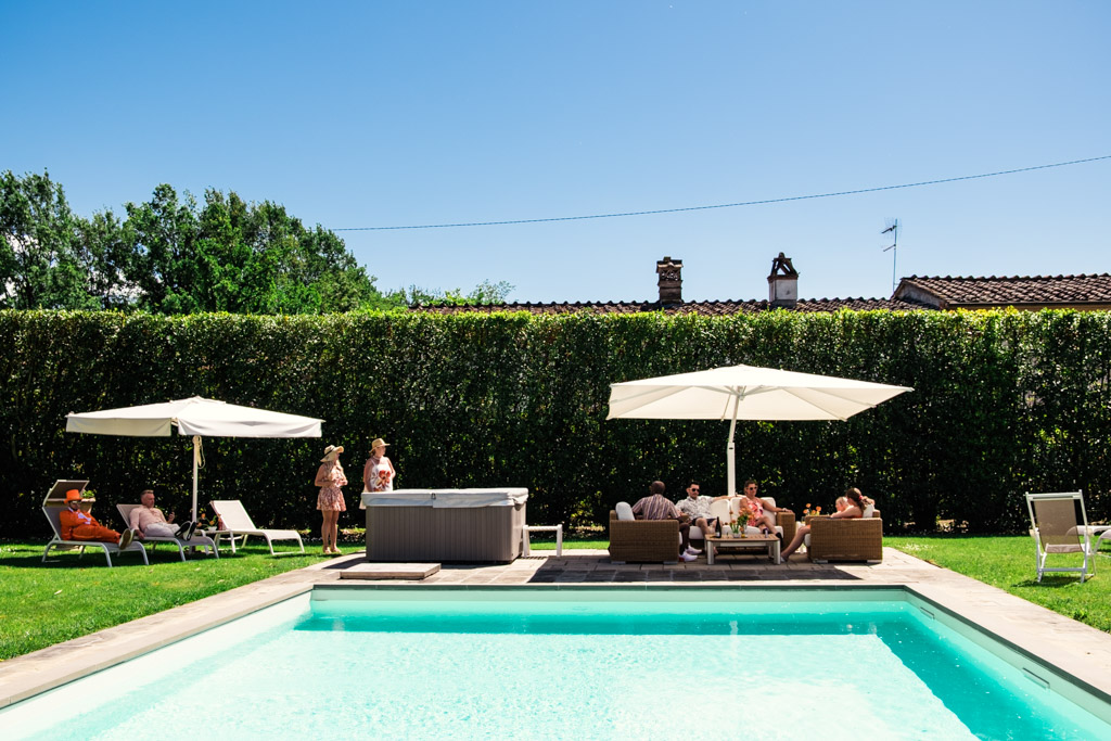 People relax by a pool, sitting under umbrellas and socializing on a sunny day during wedding pool party at Villa Moscheni in Tuscany, Italy