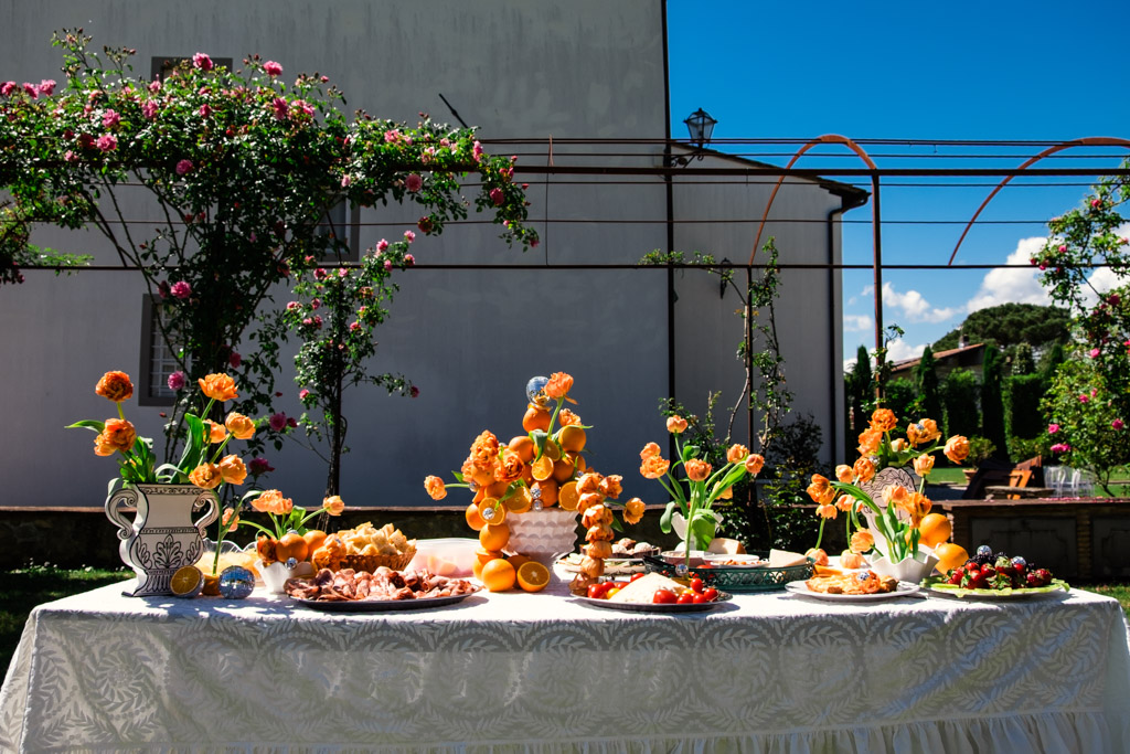 Outdoor table with orange flowers, fruit, and desserts, set against a garden and white building background for Tuscany Italy wedding weekend celebration