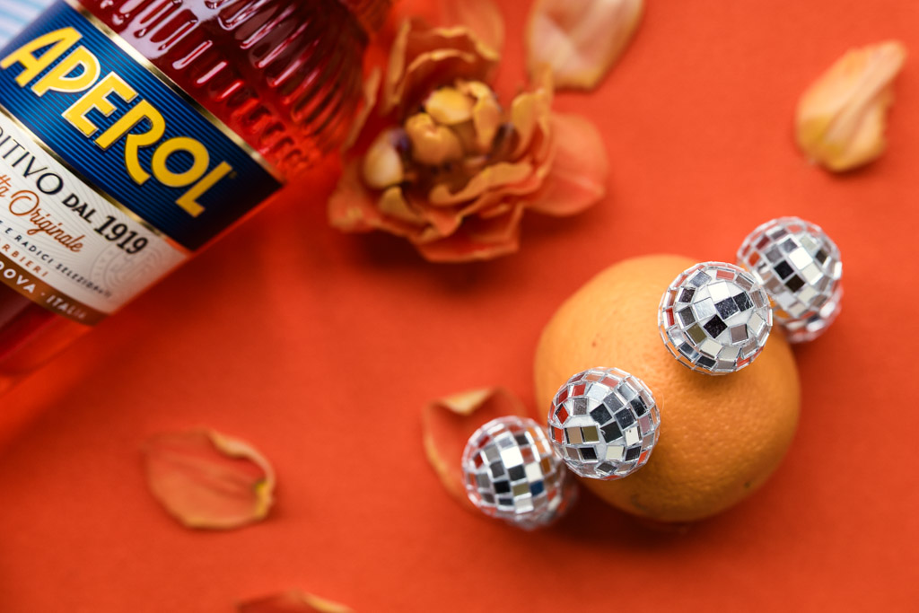 A bottle of Aperol, an orange, disco ball picks, flower, and petals on an orange background for wedding pool party at Villa Moscheni in Tuscany, Italy