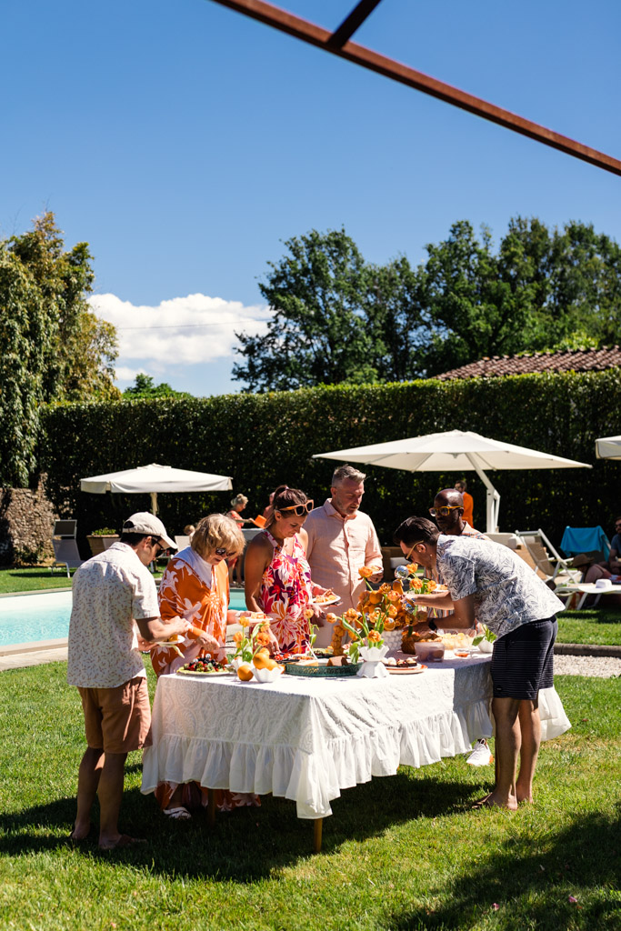 Groom and guests gather around a white table with food and flowers during poolside wedding party at Villa Moscheni in Tuscany, Italy
