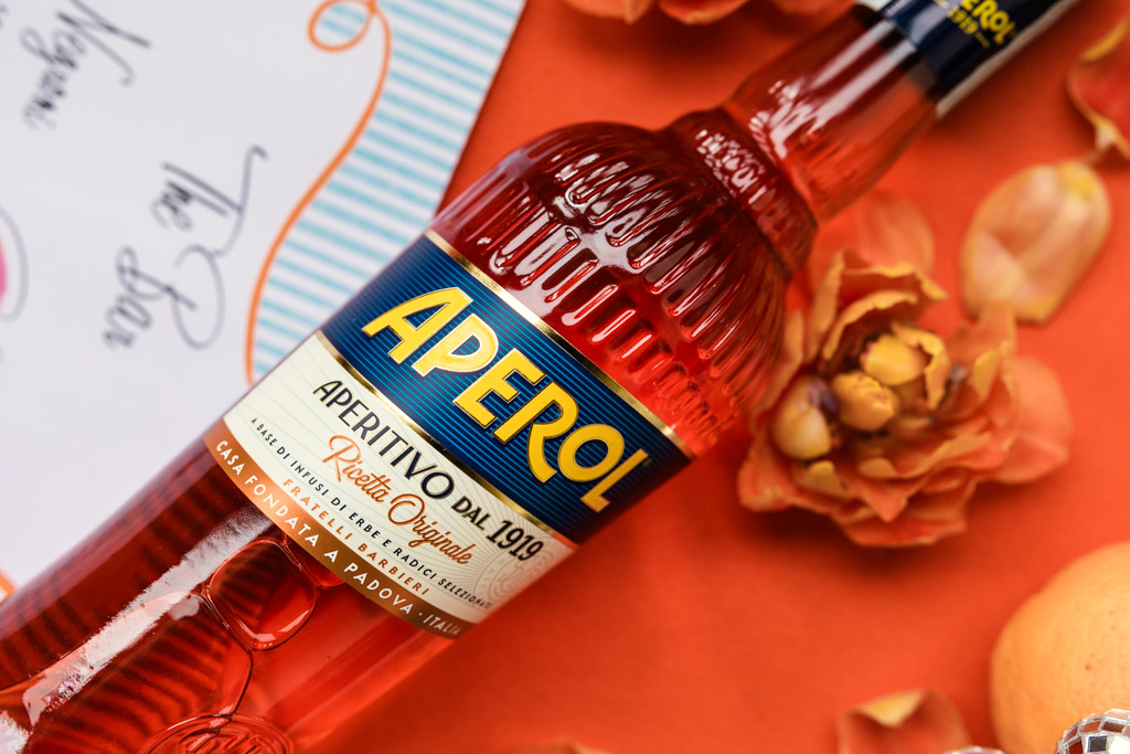 A bottle of Aperol lies on an orange surface with flowers and a card nearby for wedding pool party at Villa Moscheni in Tuscany, Italy
