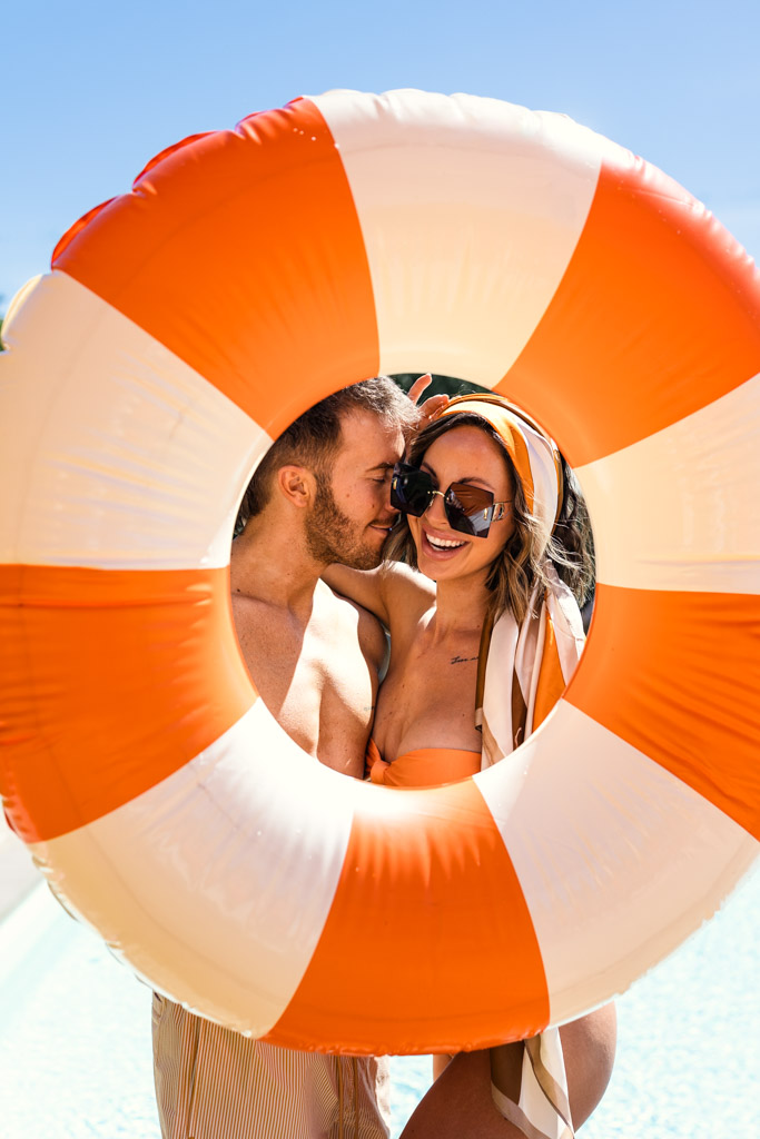 Smiling couple in swimwear pose behind an orange pool float, framed by the ring, on a sunny day during Tuscany wedding pool party
