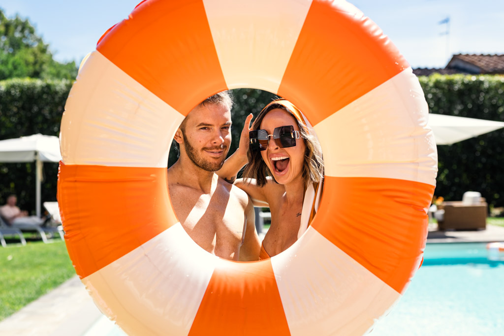 Smiling man and woman pose behind an orange pool float by a sunny outdoor swimming pool during Italy wedding weekend pool party