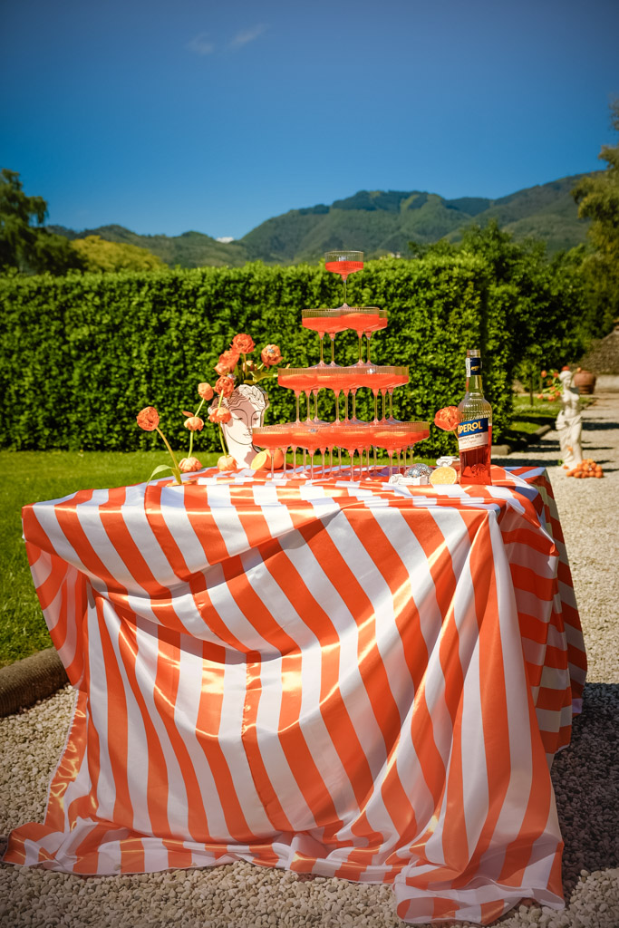 A striped table with a orange drink tower, Aperol bottle, flowers, and mountains in the background at Villa Moscheni in Tuscany, Italy