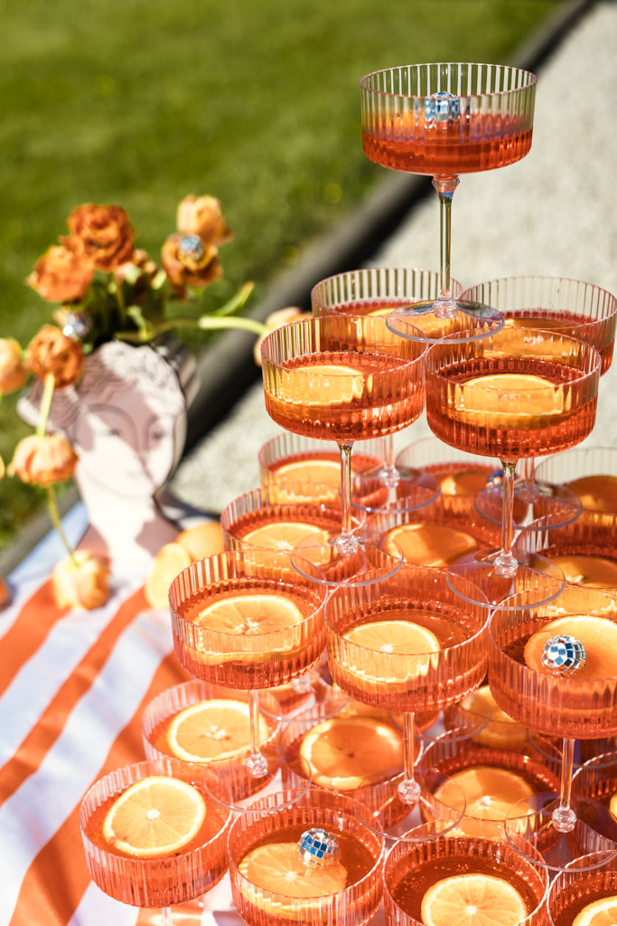 A pyramid of orange cocktails with citrus slices, set outdoors beside a vase of orange flowers for wedding pool party at Villa Moscheni in Tuscany, Italy