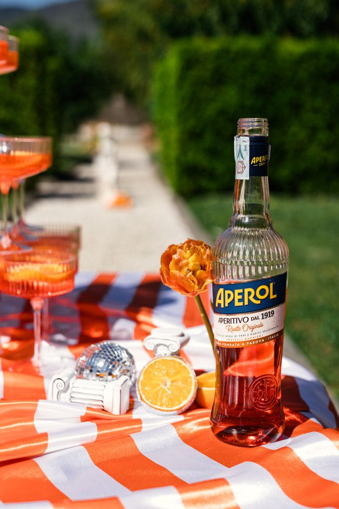 A bottle of Aperol with a flower, orange slices, and cocktails on a striped table outdoors sets the perfect scene for Italy wedding weekend pool party