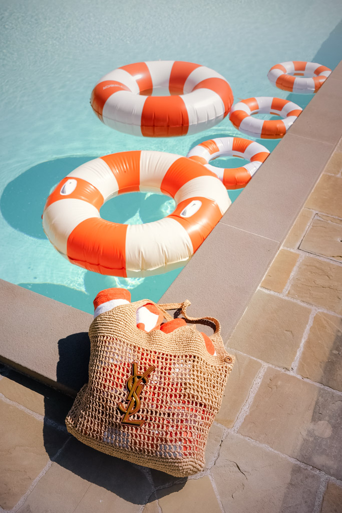 A straw YSL bag rests on poolside tiles beside orange and white pool floats in clear water, for pool party during Italy wedding weekend