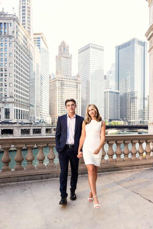 A man in a suit and a woman in a white dress hold hands, standing by a river with tall city buildings behind during their downtown Chicago summer engagement session