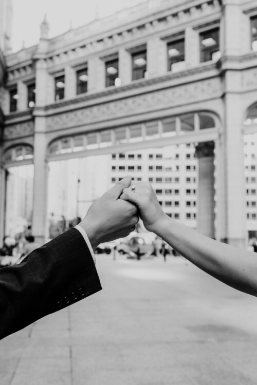 Black and white photo of two hands holding each other in front of the Wrigley building in downtown Chicago