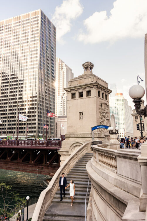 A couple walks down curved stairs near a bridge and tall buildings in bustling downtown Chicago during their summer engagement session