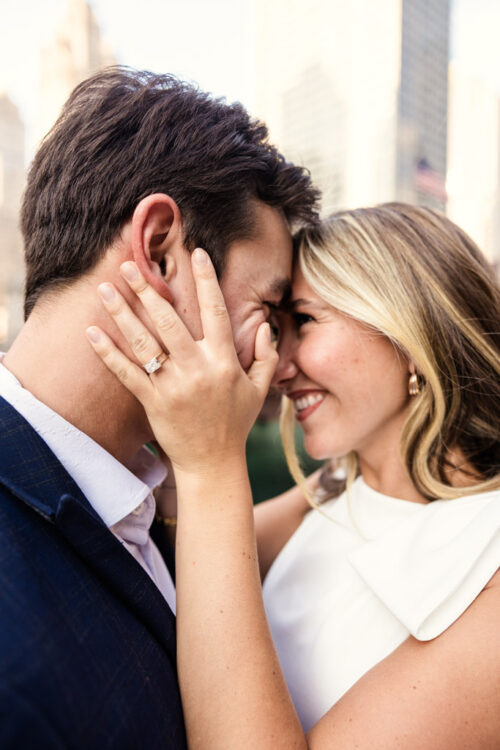 A woman smiles, touching foreheads with her fiancé during their downtown Chicago summer engagement session, proudly showing an engagement ring on her hand