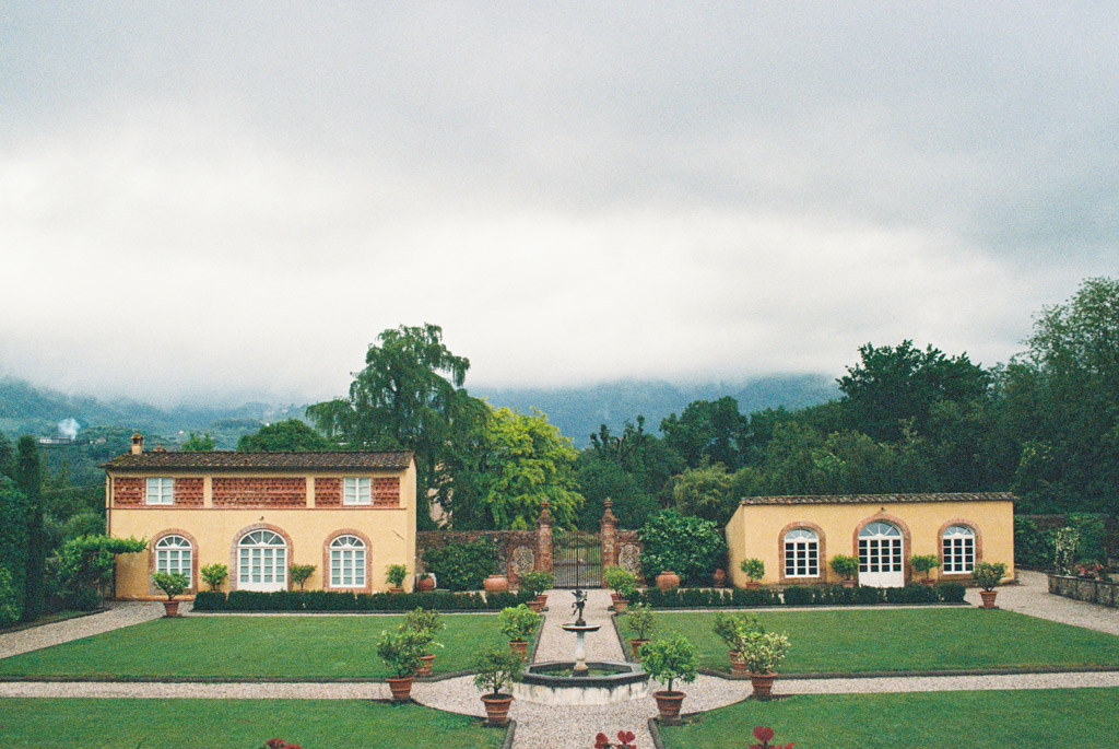 Film photo of two yellow buildings at an Italian Villa wedding face a central fountain surrounded by green lawns and potted plants, under a cloudy sky