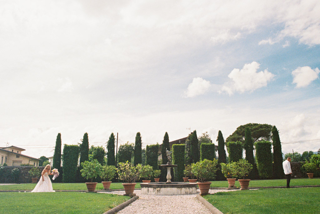 Film photo of bride in white dress holding bouquet, walking toward groom in an Italian Villa garden with tall hedges, fountain, and potted plants during their first look