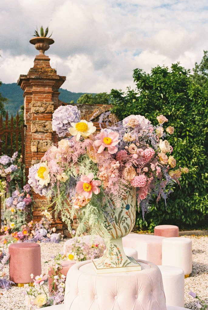 Large floral arrangement in a decorative vase outdoors, surrounded by pink and white seats and lush garden scenery for an Italian Villa wedding