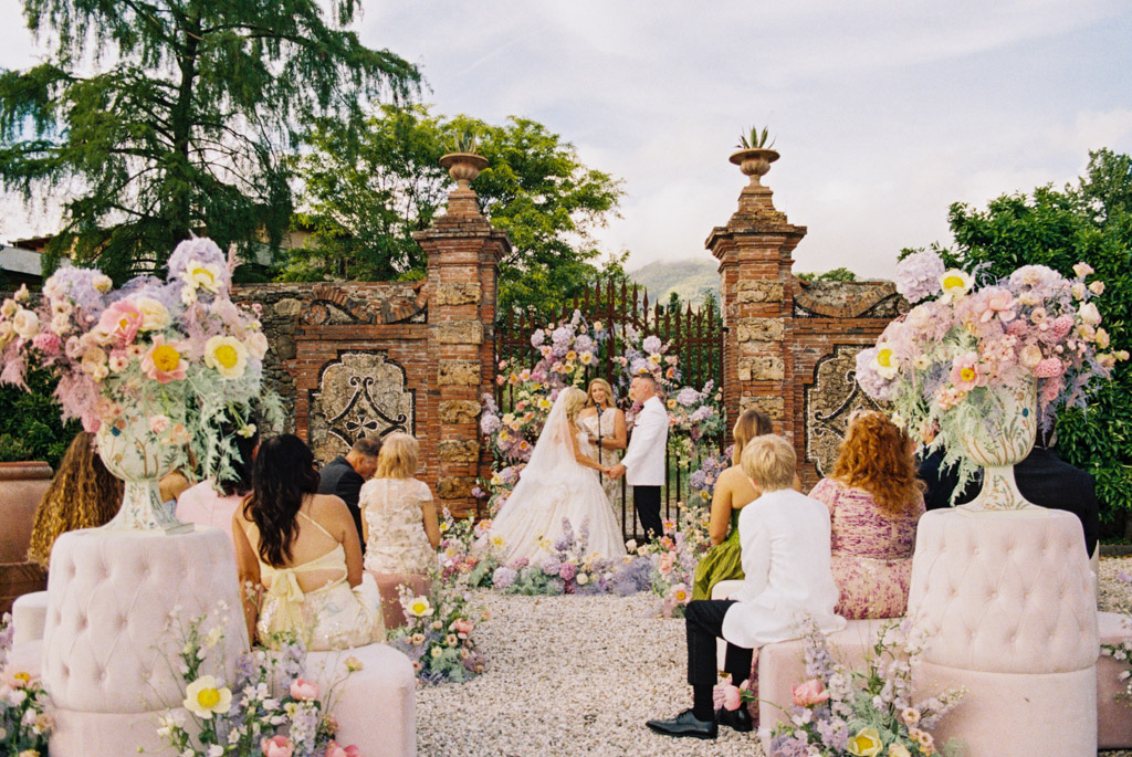 A couple weds outdoors before floral displays and guests, in front of ornate stone gates, during their Italian Villa destination wedding