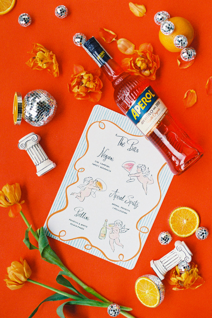 Aperol bottle, menu, oranges, flowers, disco balls, and mini columns on a bright orange surface for wedding pool party at Villa Moscheni in Tuscany, Italy