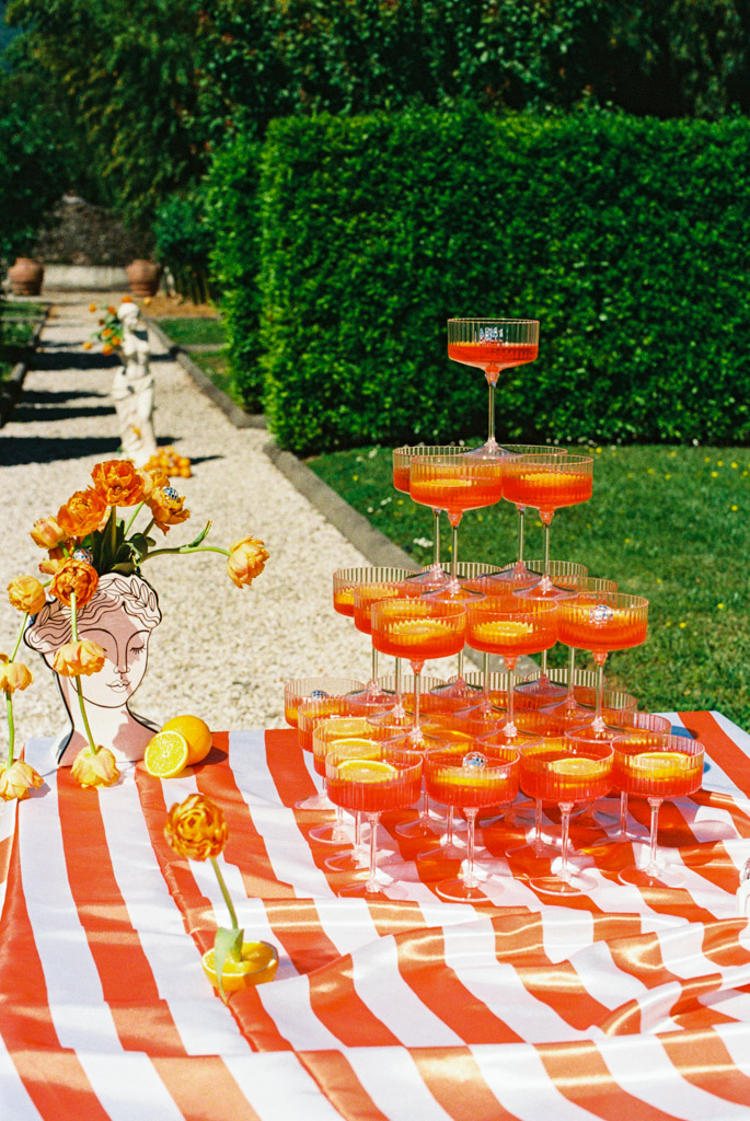 A tower of orange drinks in coupe glasses on a orange and white striped table outdoors, with flowers and citrus for post-wedding party at Villa Moscheni in Tuscany, Italy
