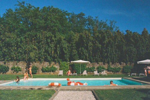 Film photo of people relaxing and swimming in a pool surrounded by greenery, lounge chairs, and umbrellas on a sunny day at Villa Moscheni in Tuscany, Italy