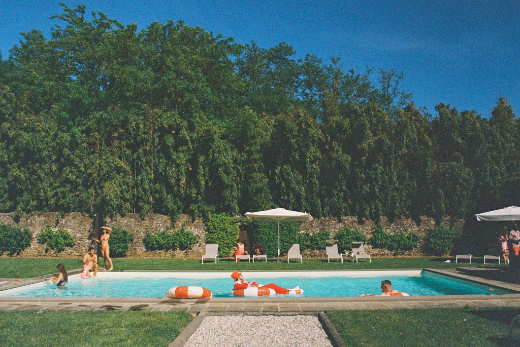 Film photo of people relaxing and swimming in a pool surrounded by greenery, lounge chairs, and umbrellas on a sunny day at Villa Moscheni in Tuscany, Italy