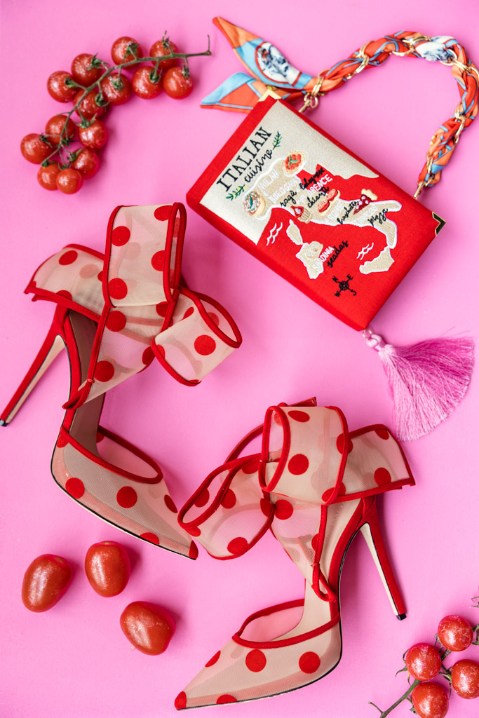 Red polka dot heels, cherry tomatoes reminiscent of a Tuscany wedding, a colorful book, and a braided necklace on a pink background.