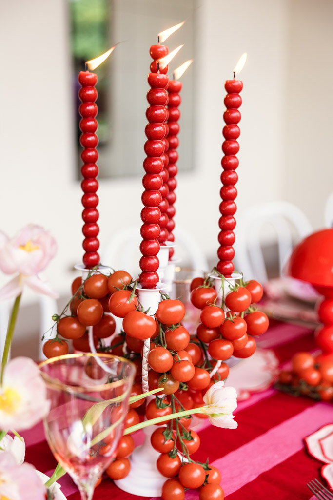 Red candles made of small spheres, decorated with clusters of cherry tomatoes on a red and pink table, evoke the vibrant charm of a Tuscany wedding.