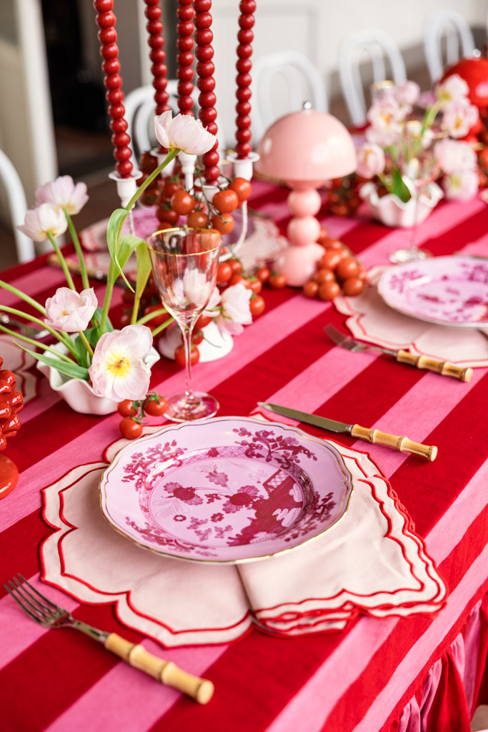 A red and pink striped table, perfect for a Tuscany wedding, with floral plates, pink tulips, red beads, and gold utensils set for a meal.