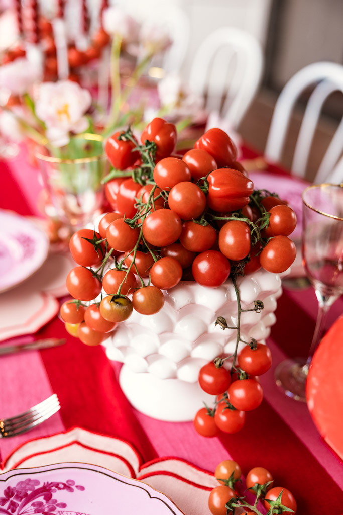 Cherry tomatoes arranged in a white textured vase evoke the rustic charm of a Tuscany wedding, set on a red and pink table setting with elegant glassware and plates.