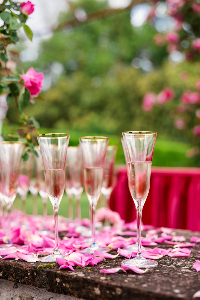 Four champagne flutes on a stone table with pink rose petals, set outdoors with flowers in the background—perfect for a romantic Tuscany wedding.