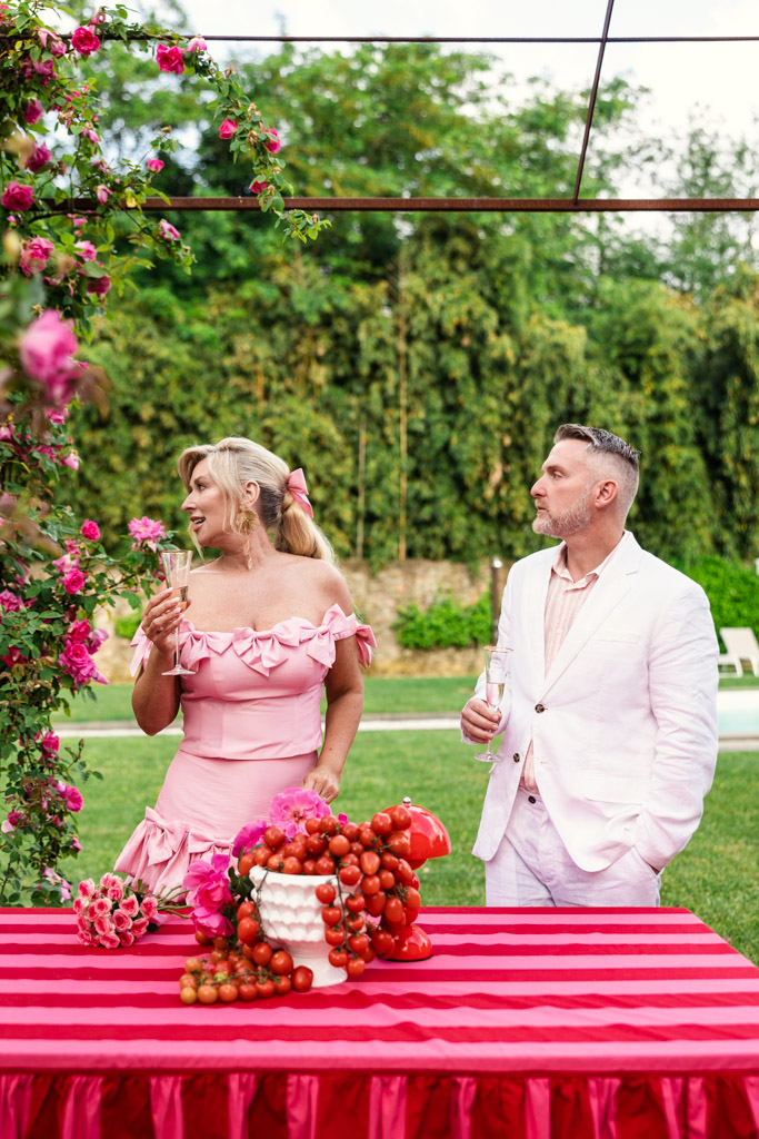 Two people in pink outfits stand by a red-striped table with fruit, surrounded by pink flowers outdoors, capturing the romantic charm of a Tuscany wedding.
