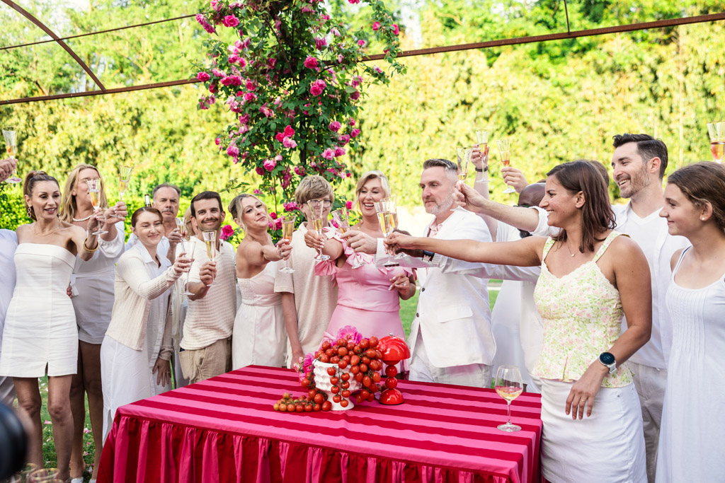 Group of people in white outfits raising glasses outdoors around a red table decorated with fruit and flowers, capturing the joyful spirit of a Tuscany wedding.